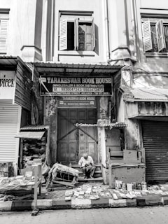 An old building facade in black and white, with open wooden shutters on the windows. Below, a bookstore named 'Tarakeswar Book House' is situated, with various books displayed on tables and the ground. A man is seated, reading a book amidst the collection. The scene appears quiet and still, capturing a moment of calm in a busy street.