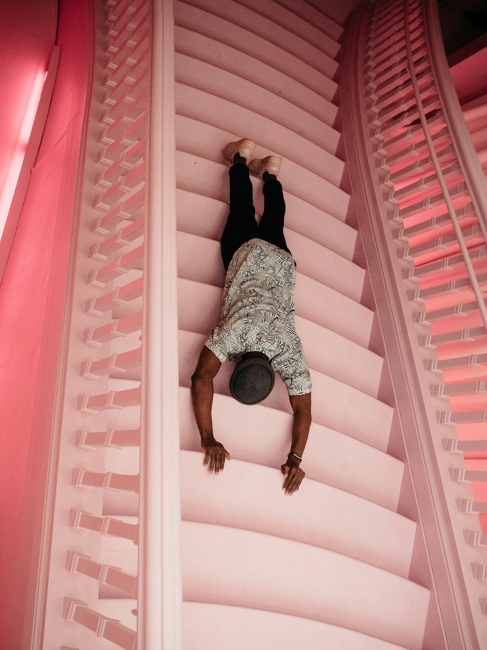 Homme vêtu d’une chemise à col rond noir et blanc et d’un pantalon noir allongé sur des escaliers en béton rose