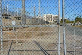 A chain-link fence with a locked gate is in the foreground, with visible padlocks securing it. Behind the fence, there is an industrial area featuring several electrical pylons and power lines. A large white building with columns is seen in the distance, situated within the complex. The ground is dry and bare with some patches of grass.