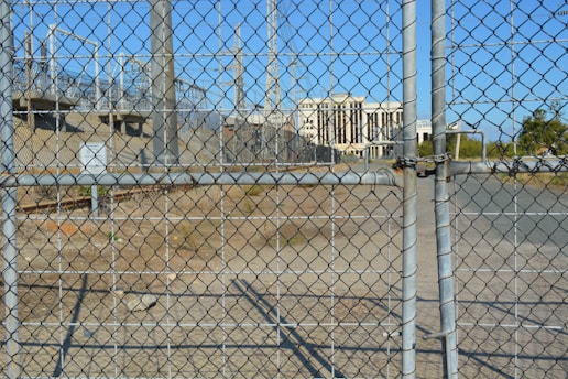 A chain-link fence with a locked gate is in the foreground, with visible padlocks securing it. Behind the fence, there is an industrial area featuring several electrical pylons and power lines. A large white building with columns is seen in the distance, situated within the complex. The ground is dry and bare with some patches of grass.