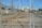 A chain-link fence with a locked gate is in the foreground, with visible padlocks securing it. Behind the fence, there is an industrial area featuring several electrical pylons and power lines. A large white building with columns is seen in the distance, situated within the complex. The ground is dry and bare with some patches of grass.