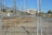 A chain-link fence with a locked gate is in the foreground, with visible padlocks securing it. Behind the fence, there is an industrial area featuring several electrical pylons and power lines. A large white building with columns is seen in the distance, situated within the complex. The ground is dry and bare with some patches of grass.
