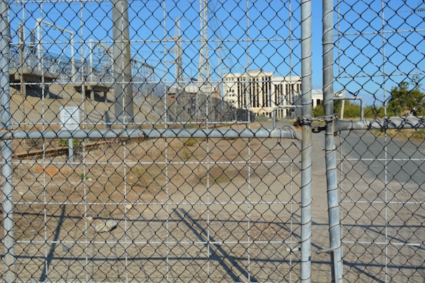 A chain-link fence with a locked gate is in the foreground, with visible padlocks securing it. Behind the fence, there is an industrial area featuring several electrical pylons and power lines. A large white building with columns is seen in the distance, situated within the complex. The ground is dry and bare with some patches of grass.