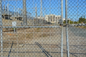 A chain-link fence with a locked gate is in the foreground, with visible padlocks securing it. Behind the fence, there is an industrial area featuring several electrical pylons and power lines. A large white building with columns is seen in the distance, situated within the complex. The ground is dry and bare with some patches of grass.