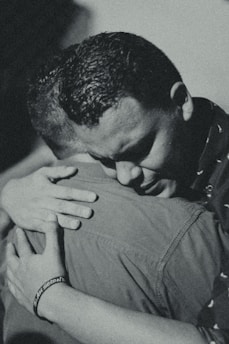 Two individuals are embracing each other closely, conveying a strong sense of comfort and support. The photograph is in black and white, highlighting the emotional intensity of the moment.