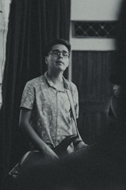 A person wearing glasses and a patterned shirt is playing a guitar. The scene is indoors with low lighting, and the ambiance is relaxed and intimate. The background features curtains and a section of a wooden wall.