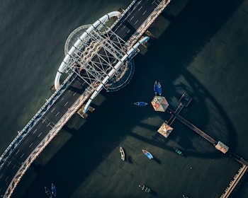 Aerial view of a bridge crossing over a body of water, with several small boats moored nearby. The bridge features a distinct architectural design with curved elements and casting shadows onto the water.