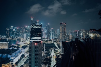 A vibrant city skyline at night, with tall skyscrapers illuminated by colorful lights. The bustling urban landscape includes a mix of modern glass structures and older buildings. A dark, cloudy sky adds depth to the scene.