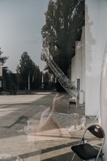 A reflection on a glass surface showcases an outdoor area with trees and a street visible in the background. Inside, there are several chairs, draped fabrics, and clear roofing sheets. The overlapping images create an ethereal and ghostly effect.