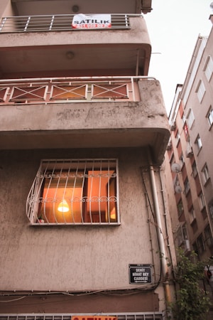 A multi-story building with a slightly aged facade. The middle floor has a window with a warm light illuminating the interior, suggesting someone is home. A sign is displayed on the balcony above, indicating the property might be for rent or sale. The building is situated on a narrow street between similar structures with visible street signage.