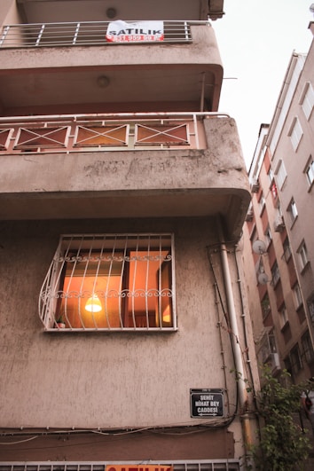 A multi-story building with a slightly aged facade. The middle floor has a window with a warm light illuminating the interior, suggesting someone is home. A sign is displayed on the balcony above, indicating the property might be for rent or sale. The building is situated on a narrow street between similar structures with visible street signage.