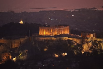 An illuminated ancient temple stands majestically atop a rocky hill against the backdrop of a dimly lit city. The temple's columns and structure are highlighted with warm, yellow lights, contrasting with the dark landscape. The distant city is barely visible under a dusky sky, creating a serene and atmospheric scene.