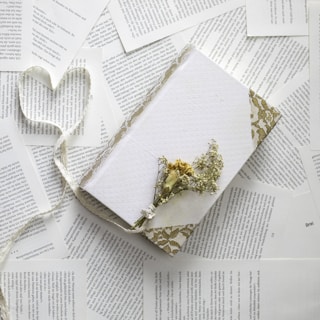 A collection of pages with text scattered around a white, decorative notebook adorned with a floral lace pattern. On top of the notebook lies a small bundle of dried flowers, and a ribbon forms the shape of a heart next to it.