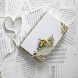 A collection of pages with text scattered around a white, decorative notebook adorned with a floral lace pattern. On top of the notebook lies a small bundle of dried flowers, and a ribbon forms the shape of a heart next to it.