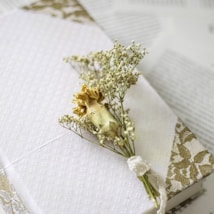 A delicate dried flower arrangement is placed on top of a white, textured fabric with a subtle floral pattern. The background includes a softly blurred texture and a lace trim detail.