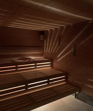 A wooden sauna with tiered seating is depicted, featuring warm lighting that casts a patterned glow on the walls and ceiling. A towel is placed on one of the benches.