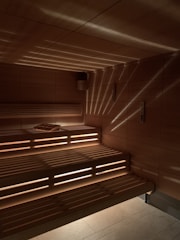 A wooden sauna with tiered seating is depicted, featuring warm lighting that casts a patterned glow on the walls and ceiling. A towel is placed on one of the benches.