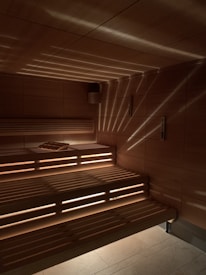 A wooden sauna with tiered seating is depicted, featuring warm lighting that casts a patterned glow on the walls and ceiling. A towel is placed on one of the benches.