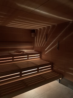 A wooden sauna with tiered seating is depicted, featuring warm lighting that casts a patterned glow on the walls and ceiling. A towel is placed on one of the benches.