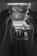 The image features a modern architectural space with large, curved walls and a glass facade overlooking a body of water. Sunlight streams through the glass, creating patterns of light and shadow on the floor. An escalator runs through the center of the space, and there are people walking in the area, engaging with the environment.