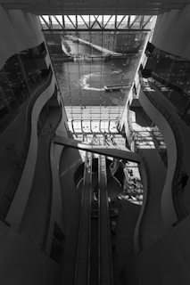 The image features a modern architectural space with large, curved walls and a glass facade overlooking a body of water. Sunlight streams through the glass, creating patterns of light and shadow on the floor. An escalator runs through the center of the space, and there are people walking in the area, engaging with the environment.