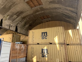 Several yellow shipping containers are stacked in a large concrete tunnel. The tunnel has a curved ceiling, and there's wood planking lining the upper sections. Sunlight partially illuminates the containers, revealing the logo 'MSC'.