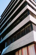 A modern building with a geometric design, featuring horizontal lines and large windows with black shutters. The structure has a sleek and minimalist appearance with concrete or stone material. Vertical blinds in various colors are visible through the lower windows.