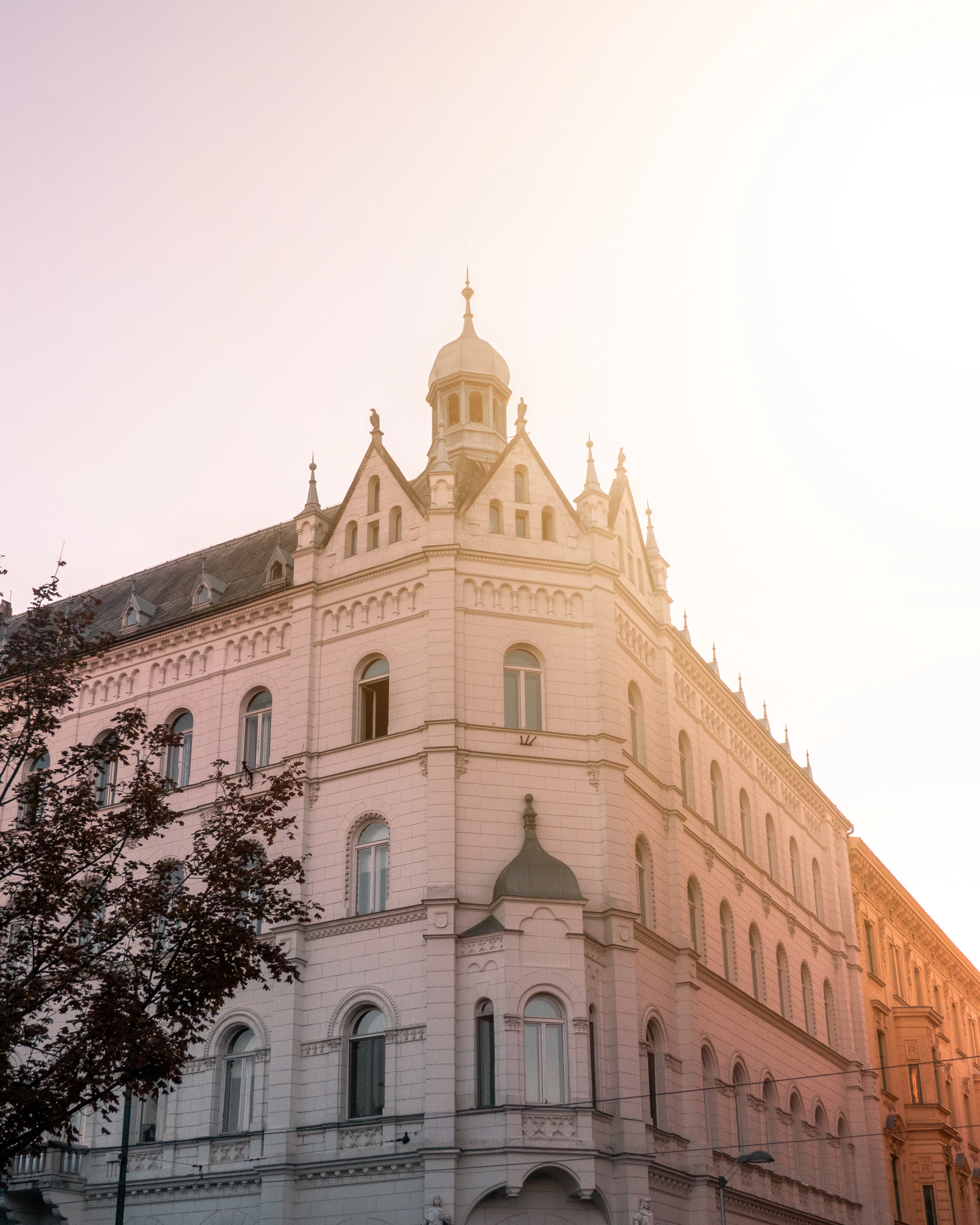 A grand pale corner building in Zagreb's Lower Town in warm hazy light, its stone facade rising through arched windows and gabled dormers to a slender cupola