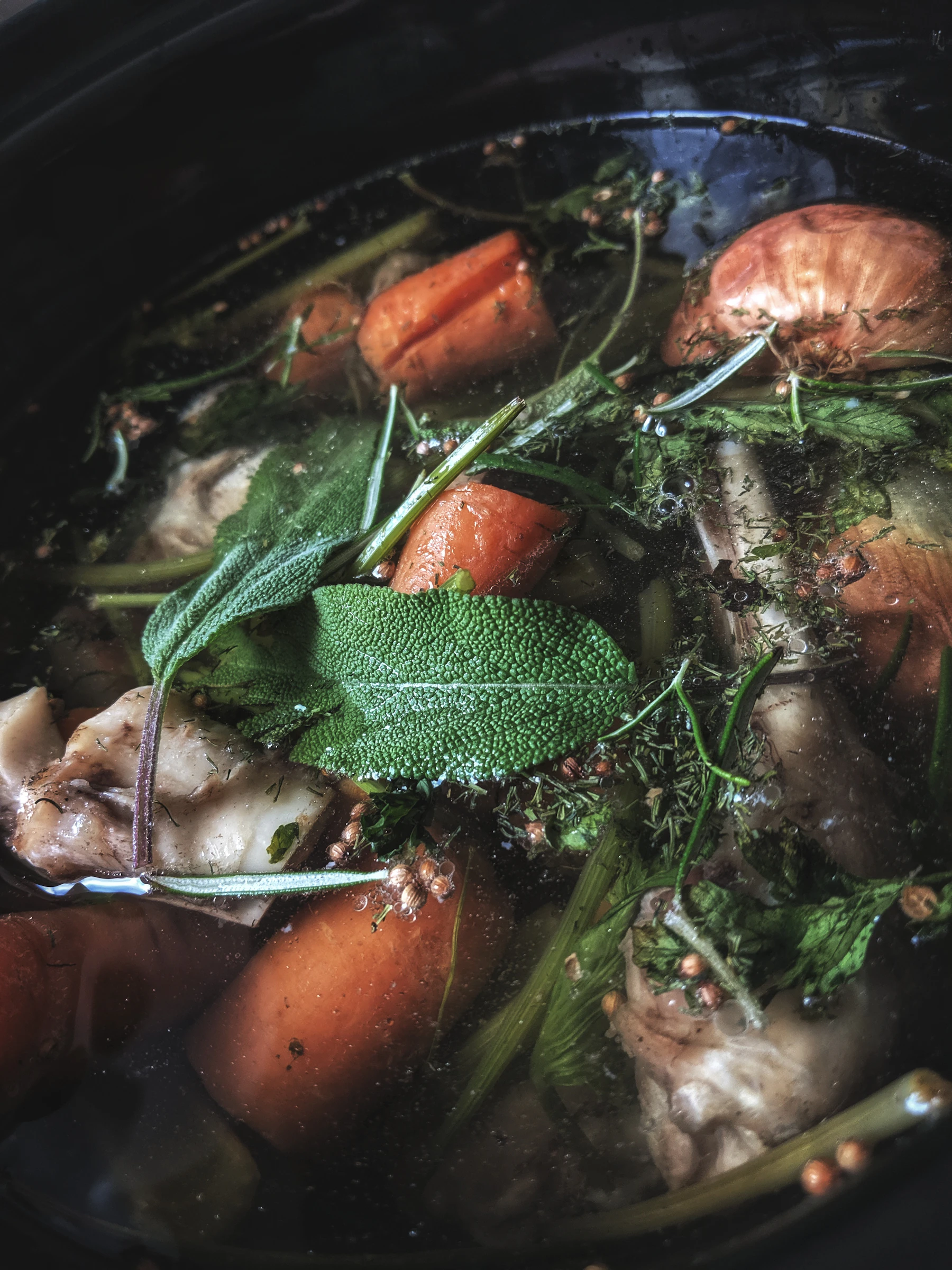 Close-up of chicken stock in the pot: chicken pieces, carrots, onion, sage leaves and whole spices under clear liquid