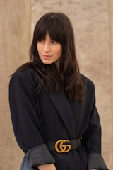 A person with long dark hair wearing a black blazer with a designer belt featuring the initials GG. The background consists of a neutral, textured wall. The person has a calm expression, looking slightly to the side.