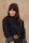 A person with long dark hair wearing a black blazer with a designer belt featuring the initials GG. The background consists of a neutral, textured wall. The person has a calm expression, looking slightly to the side.