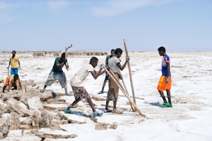 Several people stand and work on a salt flat, using tools to break and collect salt blocks. The landscape is dry and expansive, with a bright, clear sky overhead. The individuals wear casual clothing and appear to be focused on their tasks.