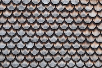 A pattern of overlapping scalloped shingles made of wood, each with a weathered and aged appearance. The shingles are arranged in rows, showcasing a texture that highlights the natural grain and color variations in the wood.