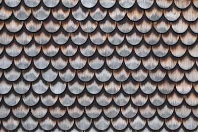 A pattern of overlapping scalloped shingles made of wood, each with a weathered and aged appearance. The shingles are arranged in rows, showcasing a texture that highlights the natural grain and color variations in the wood.