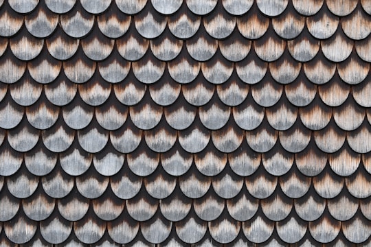 A pattern of overlapping scalloped shingles made of wood, each with a weathered and aged appearance. The shingles are arranged in rows, showcasing a texture that highlights the natural grain and color variations in the wood.