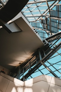 An architectural interior featuring a modern atrium with large glass panels forming part of the ceiling and walls. Metal beams and geometric patterns add a complex, structured design. Natural light streams through, casting shadows and enhancing the space's openness. Two people are visible in the lower left corner.