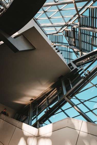 An architectural interior featuring a modern atrium with large glass panels forming part of the ceiling and walls. Metal beams and geometric patterns add a complex, structured design. Natural light streams through, casting shadows and enhancing the space's openness. Two people are visible in the lower left corner.