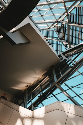 An architectural interior featuring a modern atrium with large glass panels forming part of the ceiling and walls. Metal beams and geometric patterns add a complex, structured design. Natural light streams through, casting shadows and enhancing the space's openness. Two people are visible in the lower left corner.