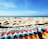 A peaceful beach setting with a clear blue sky and calm ocean waves in the background. The sandy shore is in the foreground, with a colorful, geometric-patterned beach towel partially visible.