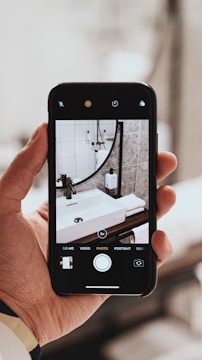 A hand holds a smartphone capturing a photo of a modern bathroom sink setup. The phone screen displays the image being taken, showing a white ceramic sink with a sleek black faucet, set against a background of white tiled and stone-textured walls. Soap dispensers and a mirror are also visible within the framed image on the phone.