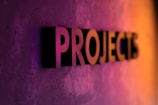 The word 'PROJECTS' appears in large, bold, three-dimensional letters against a textured surface, with vibrant lighting that transitions from purple to orange, creating a dynamic gradient effect.