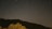 A dark night sky with visible star trails indicating long exposure. Below, a silhouette of a mountain range and a tree in the foreground.