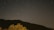 A dark night sky with visible star trails indicating long exposure. Below, a silhouette of a mountain range and a tree in the foreground.