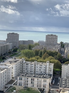 Several apartment buildings are visible in a coastal cityscape. In the background, the sea stretches beneath a cloudy sky. Green trees and a few parked cars can be seen between the structures.