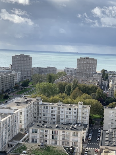 Several apartment buildings are visible in a coastal cityscape. In the background, the sea stretches beneath a cloudy sky. Green trees and a few parked cars can be seen between the structures.