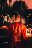 A person wearing a richly decorated red robe with intricate embroidery stands with their back to the viewer. The scene is softly illuminated by warm, glowing lights from lanterns resembling lotus flowers. The background features blurred, dark silhouettes of trees, creating a mystical ambiance.