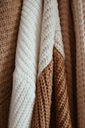 Close-up view of several knitted fabrics in beige, cream, and light brown tones. The fabrics display various textures and patterns, with prominent stitches and a cozy appearance.