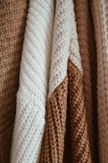 Close-up view of several knitted fabrics in beige, cream, and light brown tones. The fabrics display various textures and patterns, with prominent stitches and a cozy appearance.