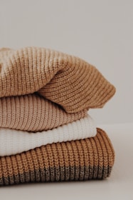 A neatly stacked pile of knitted sweaters in various shades, including cream, tan, and brown. The texture of the fabric is clearly visible, suggesting warmth and coziness.