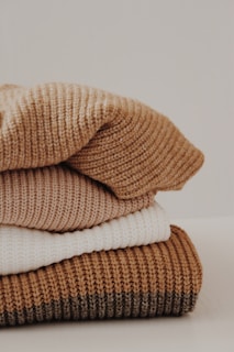 A neatly stacked pile of knitted sweaters in various shades, including cream, tan, and brown. The texture of the fabric is clearly visible, suggesting warmth and coziness.